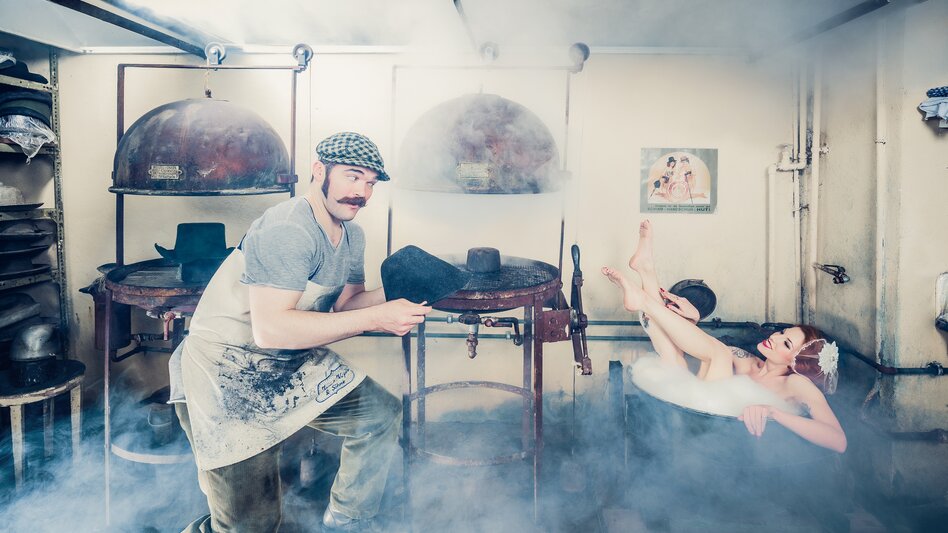 In der nebeligen Hutwerkstatt ist eine alte Badewanne aufgestellt, in der eine Dame badet. Der Hutmacher bietet ihr einen neu angefertigten Hut an.  | © Karin Krahl-Wichmann