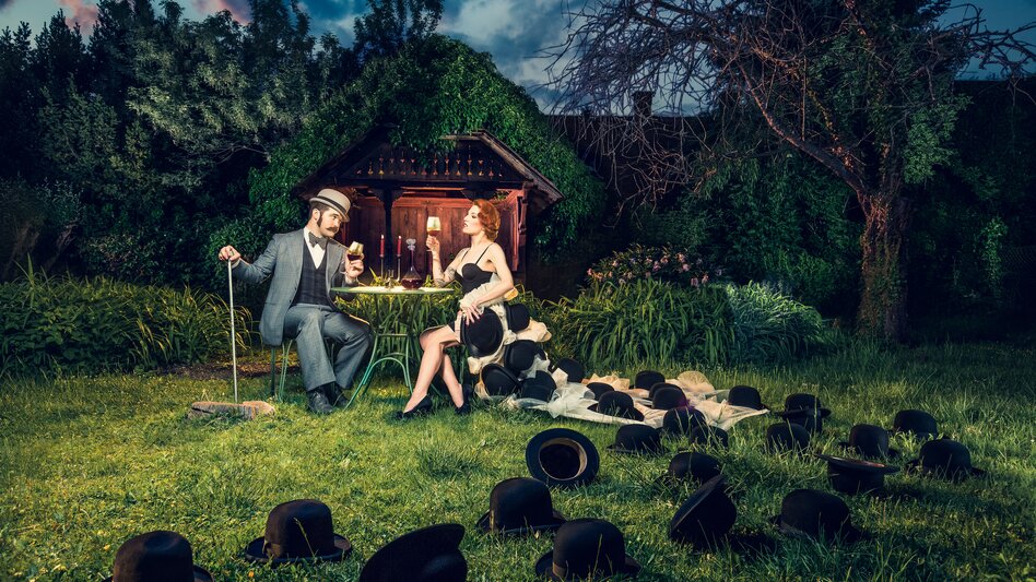 Ein Paar sitzt beim Abendessen im Grünen. Auf der Wiese rundherum liegen schwarze Hüte. | © Karin Krahl-Wichmann