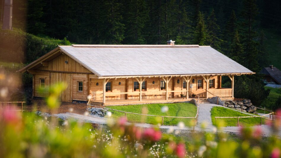 Die Latschenbrennerei, ein Gebäude aus hellem Holz, steht umringt von zarten Wiesenblumen inmitten der Wiesen. | © UMJ