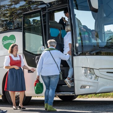 Die Busreisenden steigen in den weißen Reisebus ein. Die Reiseleitung im Dirndl begrüßt die Einsteigenden mit einem herzlichen Lächeln. | © Busreisen Steiermark