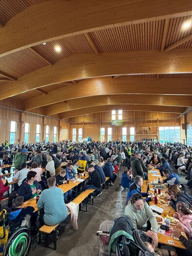 A large crowd in a cozy hall with wooden walls. The guests are sitting at long tables and enjoying their food. | © SOMA