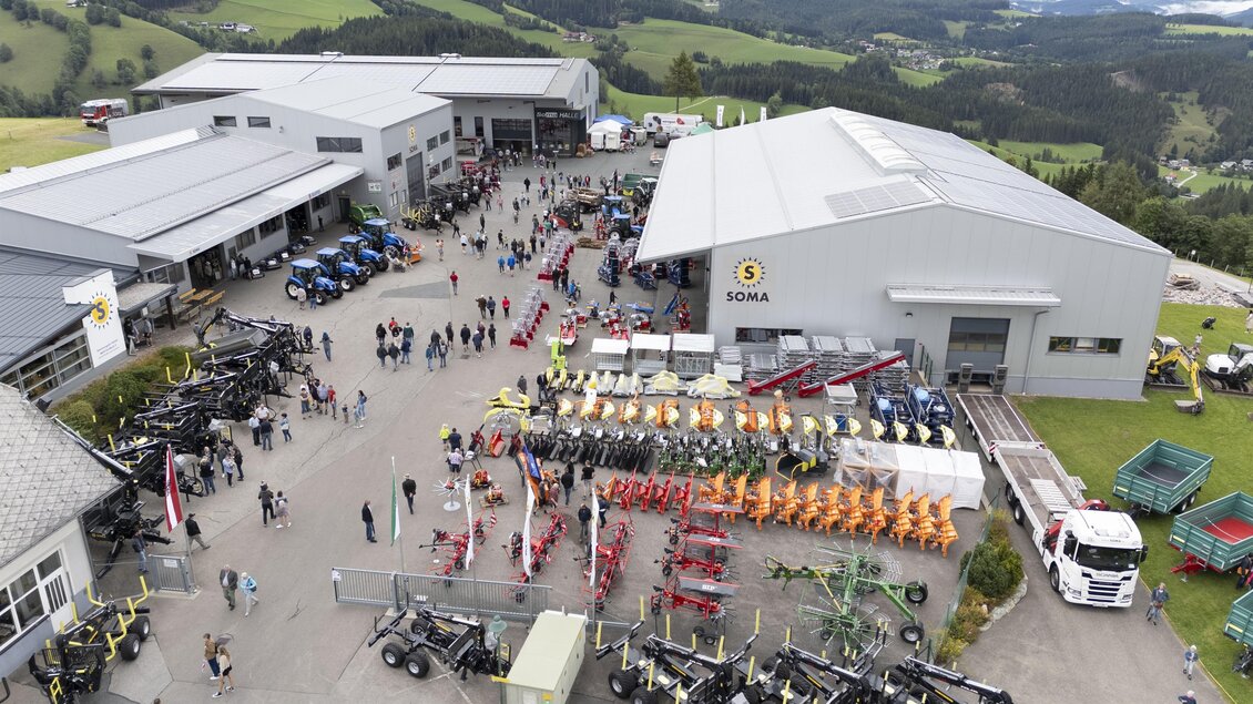Ein großer Betriebshof mit vielen Besuchern und landwirtschaftlichen Maschinen ist zu sehen. Im Hintergrund gibt es grüne Hügel und Gebäude. | © SOMA