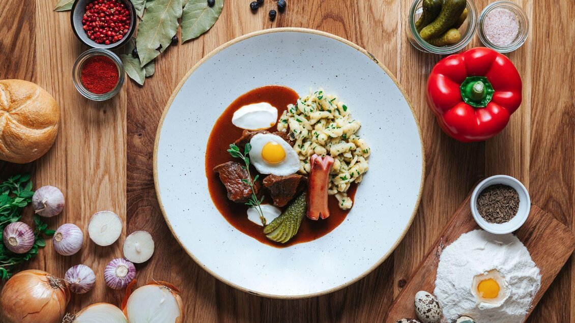 Ein Teller mit herzhafter Speise, bestehend aus Fleisch, Spätzle und einem weichgekochten Ei. Um den Teller sind verschiedene Zutaten wie Gemüse und Gewürze angeordnet. | © Karl Steinegger