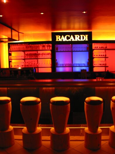 A stylish bar with illuminated shelves and colorful lights. The bar stools are arranged in a modern and inviting way. | © Hotel Seebacherhof