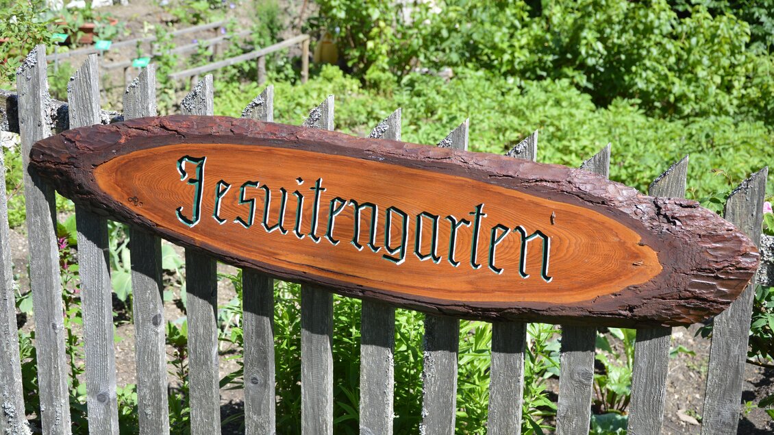 Ein Holzschild mit der Aufschrift "Jesuitengarten" steht an einem Gartenzaun. Im Hintergrund sind grüne Pflanzen zu sehen. | © Naturpark Sölktäler