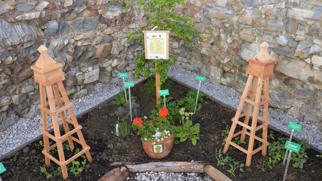 Ein kleiner Garten mit zwei hölzernen Stelen und einer Blumentopfmitte. Die Wände bestehen aus Steinen, und es wachsen verschiedene Pflanzen. | © Naturpark Sölktäler