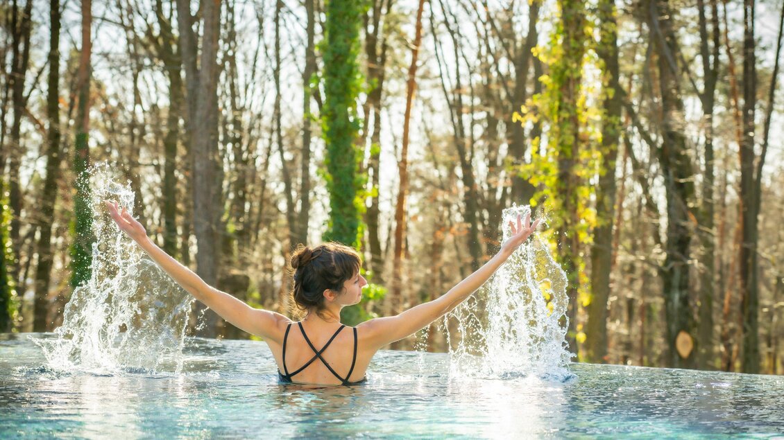 Eine Frau steht im Wasser und genießt die sprudelnden Wasserfontänen. Im Hintergrund sind Bäume und natürliche Vegetation zu sehen. | © RETTER Bio- Natur- Resort
