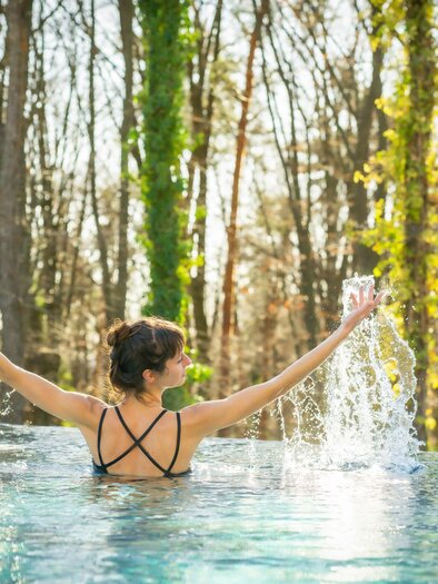 Eine Frau steht im Wasser und genießt die sprudelnden Wasserfontänen. Im Hintergrund sind Bäume und natürliche Vegetation zu sehen. | © RETTER Bio- Natur- Resort