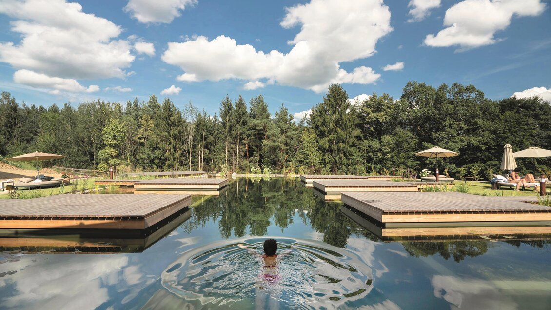 Eine ruhige Umgebung mit einem klaren Wasserbecken und Wolken im blauen Himmel. Im Wasser ist eine Person, die Wellensprünge macht. | © RETTER Bio- Natur- Resort