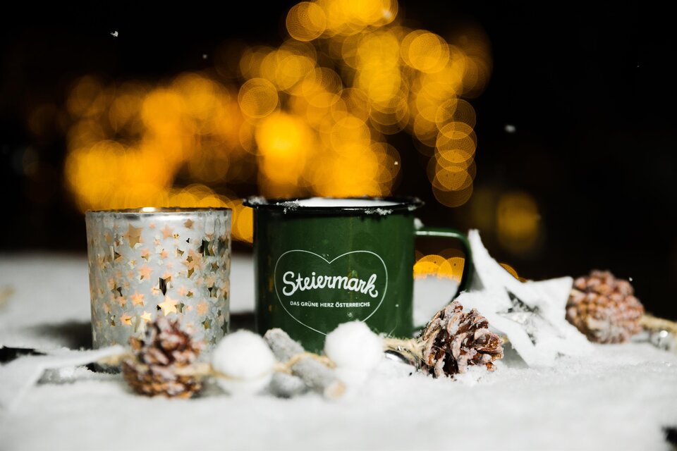 Eine grüne Tasse mit der Aufschrift „Steiermark“ steht im Schnee, neben einem dekorativen Glas. Im Hintergrund strahlen warme Lichter und eine winterliche Atmosphäre. | © Lupi Spuma