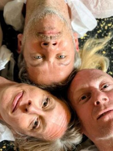 Drei Personen liegen auf dem Boden und schauen zur Kamera. Sie tragen helle Oberbekleidung und der Hintergrund hat ein auffälliges Muster. | © Theaterhaus Graz