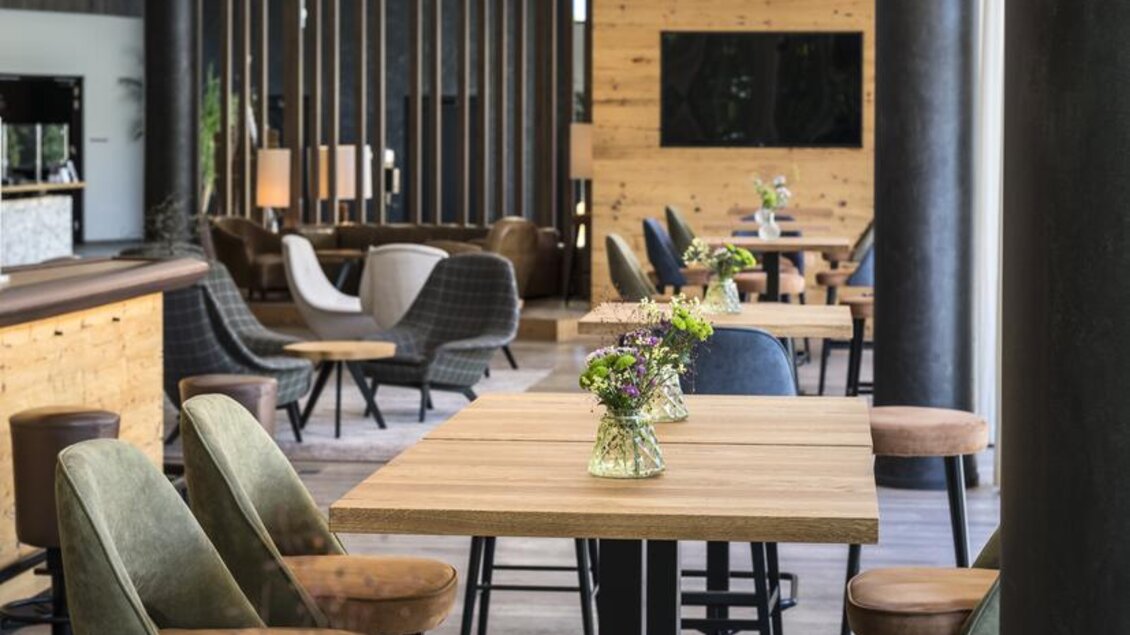Ein modern gestalteter Innenraum mit Holztheke und stilvollen Sitzplätzen. Auf den Tischen stehen kleine Vasen mit frischen Blumen. | © Falkensteiner Hotel Schladming/ Fotograf Walter Luttenberger