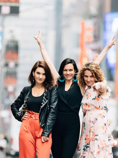 Three women are happily posing on a busy street. The city atmosphere and colorful advertisements in the background give the picture a lively mood. | © helgaTraxler