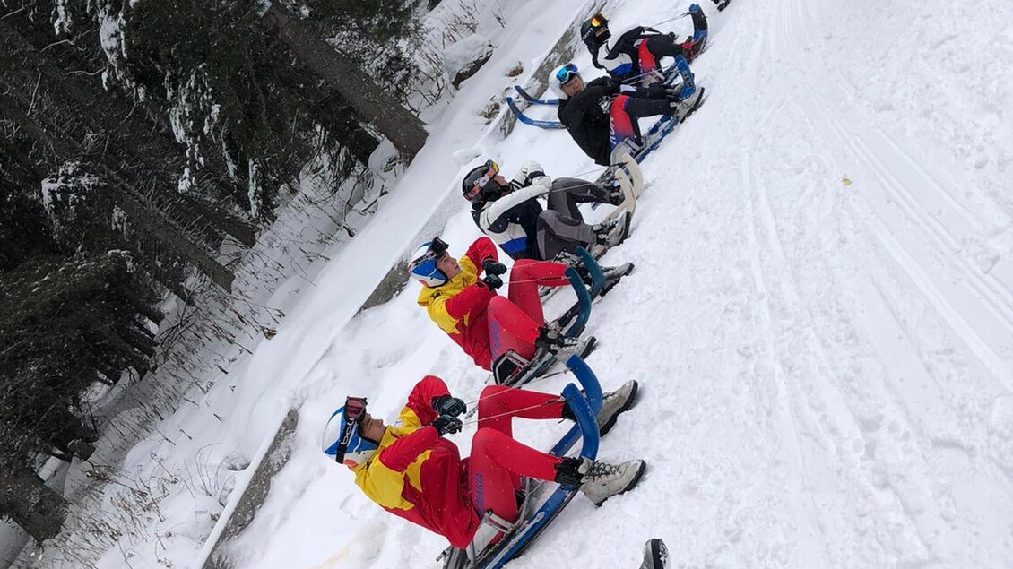 Eine Gruppe von Personen sitzt auf Schlitten im Schnee. Im Hintergrund ist ein Trainer zu sehen, der Anweisungen gibt. | © Rennrodelverein