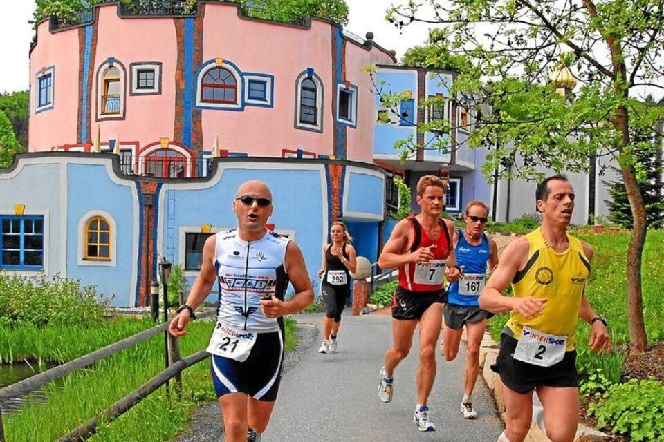 Eine Gruppe von Läufern bewegt sich entlang eines Pfades in einer malerischen Umgebung. Im Hintergrund ist ein buntes, einzigartiges Gebäude zu sehen.