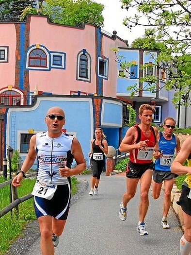 A group of runners is moving along a path in a picturesque setting. In the background, a colorful, unique building can be seen.