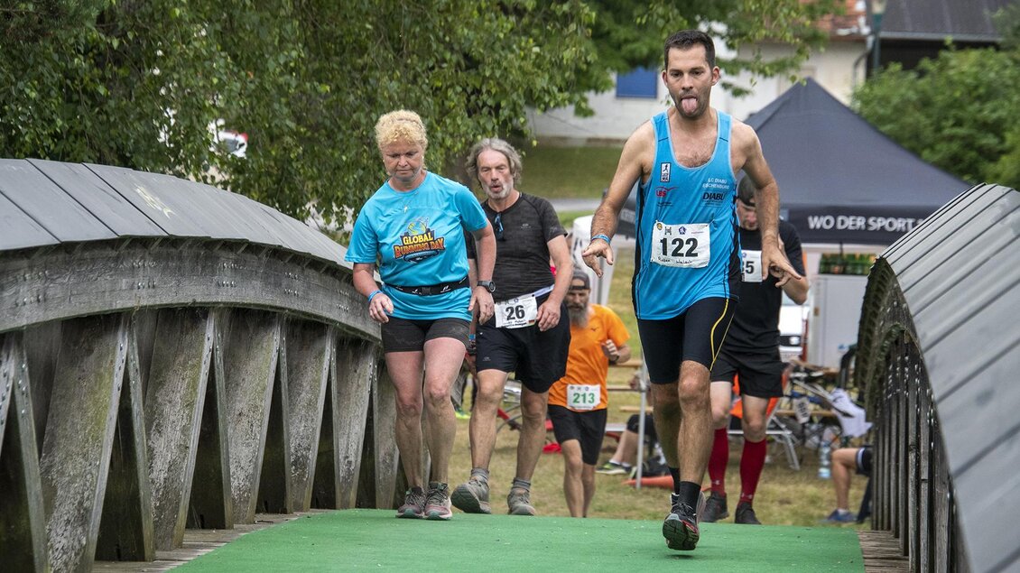 Eine Gruppe von Läufern überquert eine Brücke auf einem Wettkampf. Im Hintergrund sind Bäume und ein Veranstaltungszelt sichtbar. | © Christian Thomaser