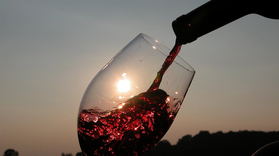 Ein Glas Rotwein wird aus einer Flasche eingeschenkt. Im Hintergrund ist die Sonne zu sehen, die bei Sonnenuntergang scheint. | © @Josi Thaller