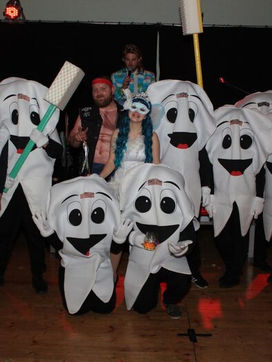 A group of people in tooth-themed costumes, including a toothbrush and a tube of toothpaste. They are happily posing on a stage with lights in the background. | © Freizeitclub Sebersdorf