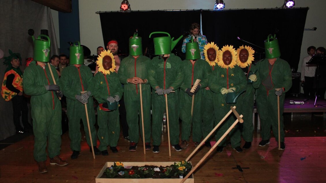 Eine Gruppe von Menschen steht in grünen Kostümen mit Gießkanne- und Sonnenblumenmasken. Sie halten Werkzeuge und stehen vor einer Wagen mit Blumen. | © Freizeitclub Sebersdorf