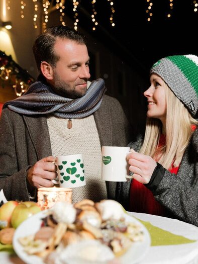A couple stands at a table, holding cups in their hands and smiling at each other. In the background, festive lights and a stall can be seen. | © Schilcherland Steiermark
