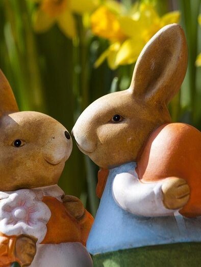 Two cute Easter bunny figures face each other. In the background, yellow daffodils are blooming. | © Pixabay