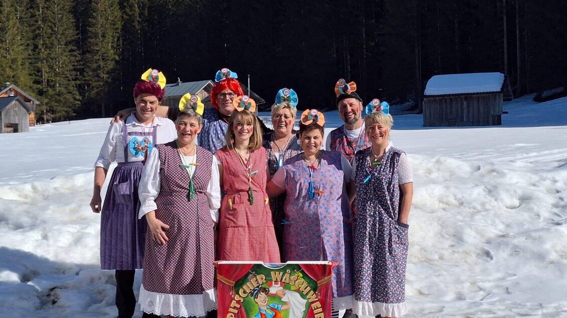 Eine Gruppe von Leuten in traditionellen Kostümen steht im Schnee. Sie tragen bunte Haarnetze und lächeln vor einer Winterlandschaft. | © Martina Gaisberger