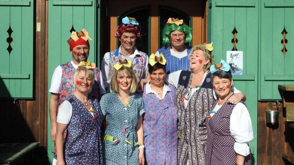 Eine fröhliche Gruppe von acht Personen in bunten Kostümen mit lustigen Hüten. Sie stehen vor einer Holztür mit grünen Fensterläden und lächeln in die Kamera. | © Martina Gaisberger
