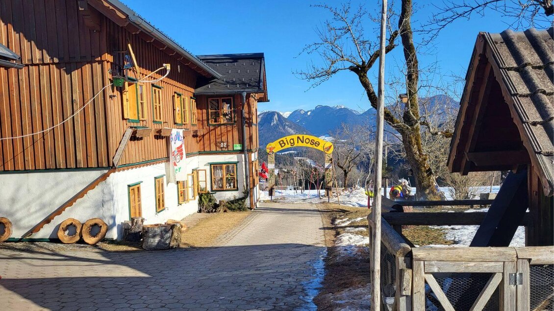 Ein traditionelles Holzhaus mit einem hellen blauen Himmel und schneebedeckten Bergen im Hintergrund. Ein buntes Schild mit der Aufschrift „Big Nose“ ist gut sichtbar. | © Petra Kirchschlager