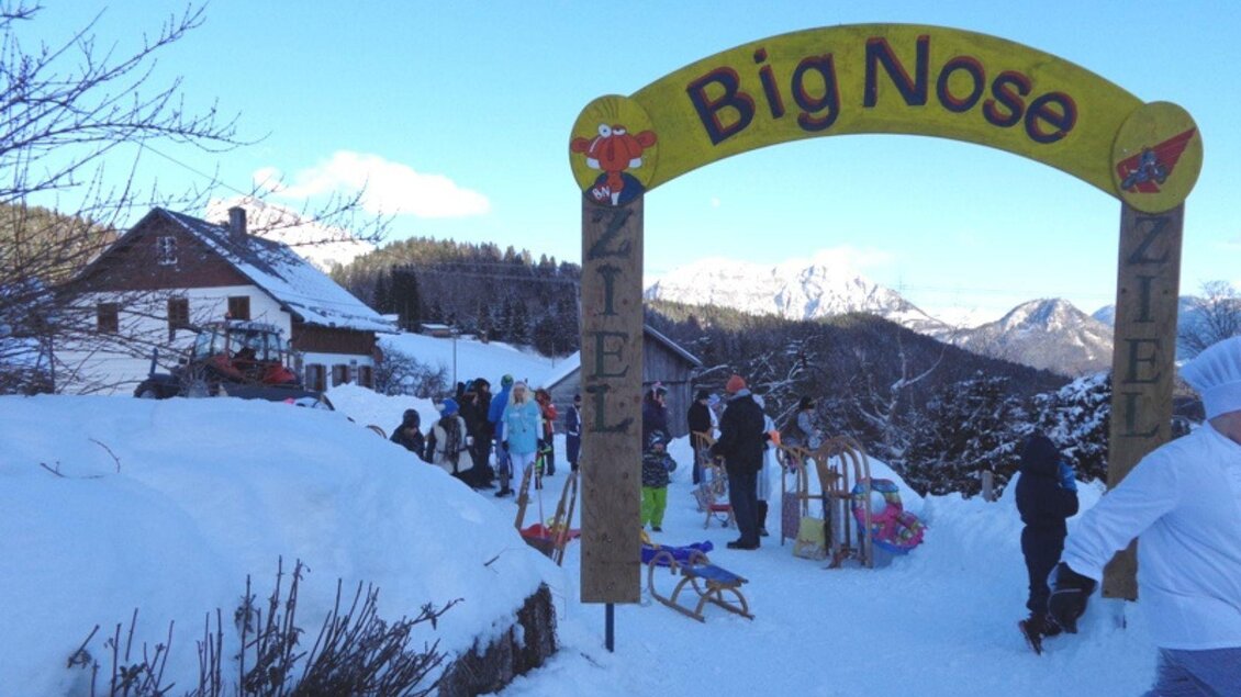 Ein festlicher Eingang mit dem Schriftzug "Big Nose" in einer schneebedeckten Umgebung. Viele Menschen genießen Aktivitäten im Schnee vor einem traditionellen Haus. | © Kirchschlager Petra