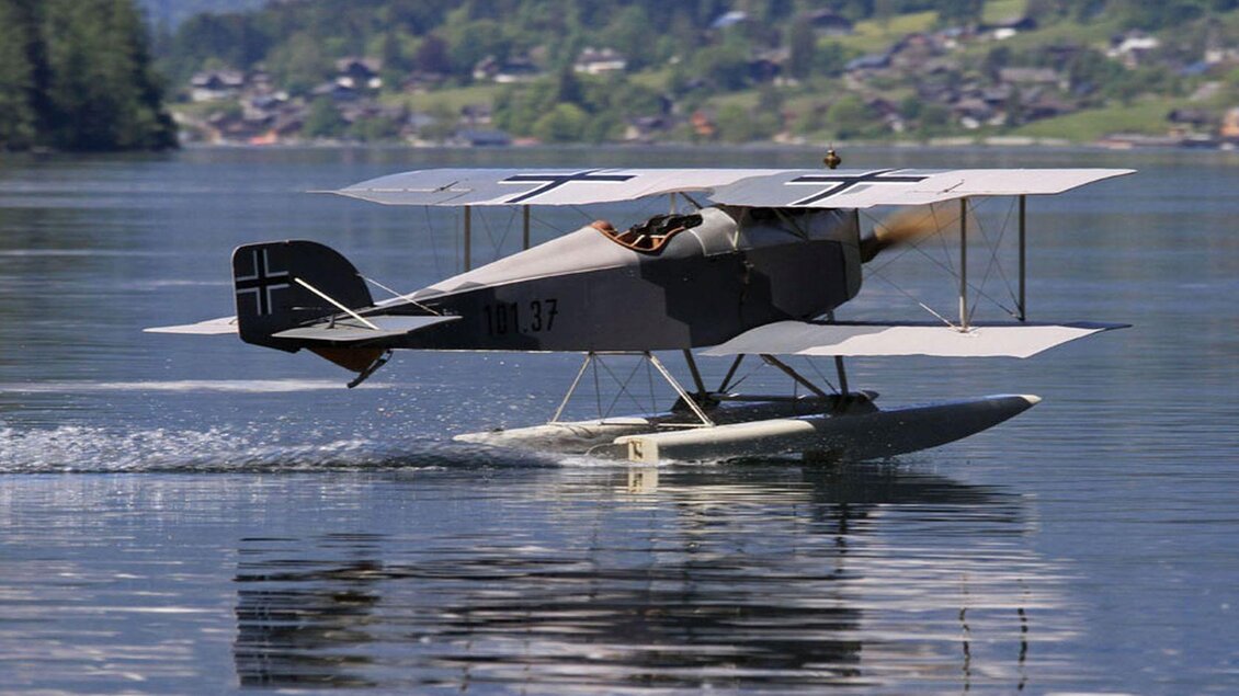 Ein historisches Flugzeug landet auf einem ruhigen Gewässer. Im Hintergrund sind grüne Hügel und eine kleine Ortschaft sichtbar. | © Ian Walter