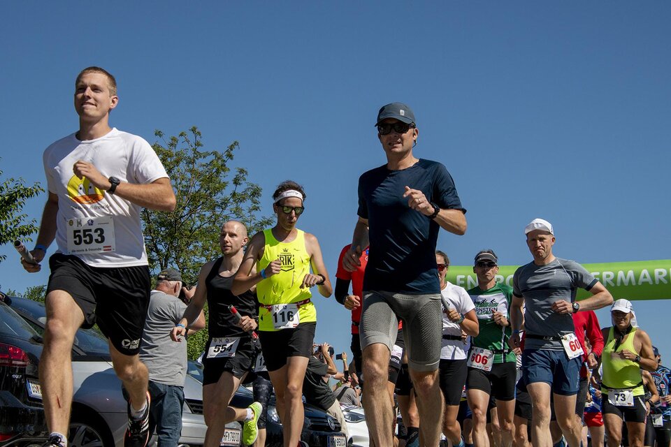 Eine Gruppe von Läufern joggt unter klarem blauen Himmel. Im Hintergrund sind Bäume und eine Startlinie zu sehen. | © Christian Thomaser