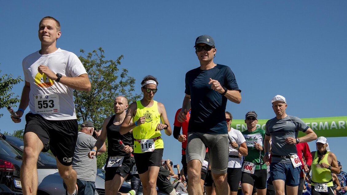 Eine Gruppe von Läufern joggt unter klarem blauen Himmel. Im Hintergrund sind Bäume und eine Startlinie zu sehen. | © Christian Thomaser