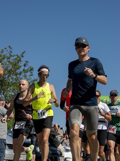 Eine Gruppe von Läufern joggt unter klarem blauen Himmel. Im Hintergrund sind Bäume und eine Startlinie zu sehen. | © Christian Thomaser