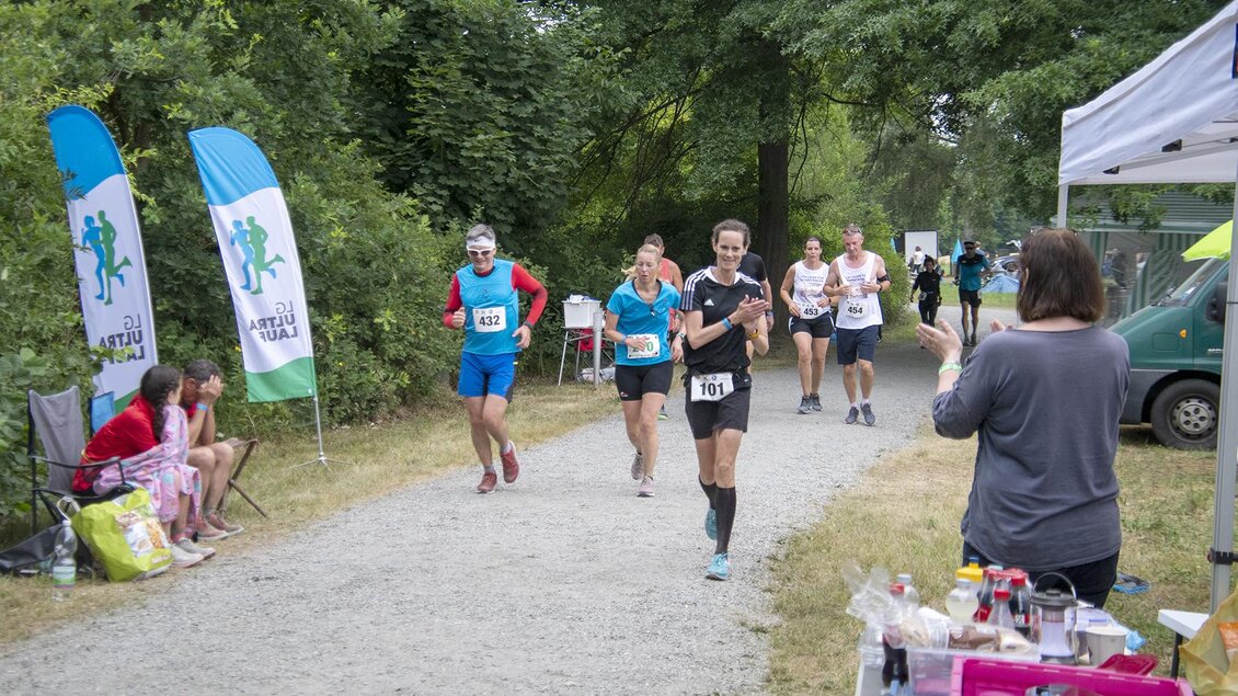 Eine Gruppe von Läufern nimmt an einem Wettlauf auf einem Kiesweg teil. Im Hintergrund befinden sich Zuschauer und ein Stand mit Getränken. | © Christian Thomaser