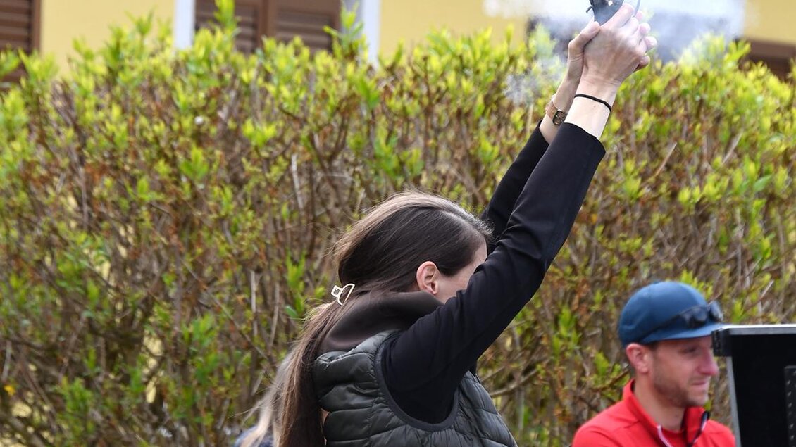 Eine Zuschauerin hebt eine Handykamera in die Luft, während sie ein Feuerwerk oder ein Signal abfeuert. Im Hintergrund sind farbige Pflanzen und Personen zu sehen. | © Christian Thomaser