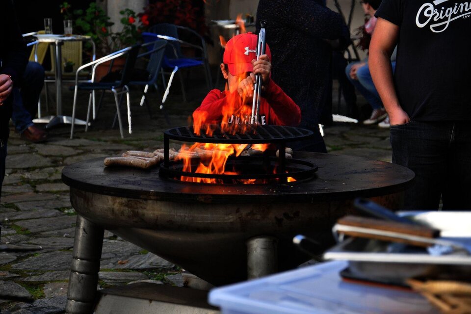 Ein Kind in rotem Hut steht an einem Grill, während Flammen aufsteigen. Um ihn herum sind einige Menschen und Tische im Freien.