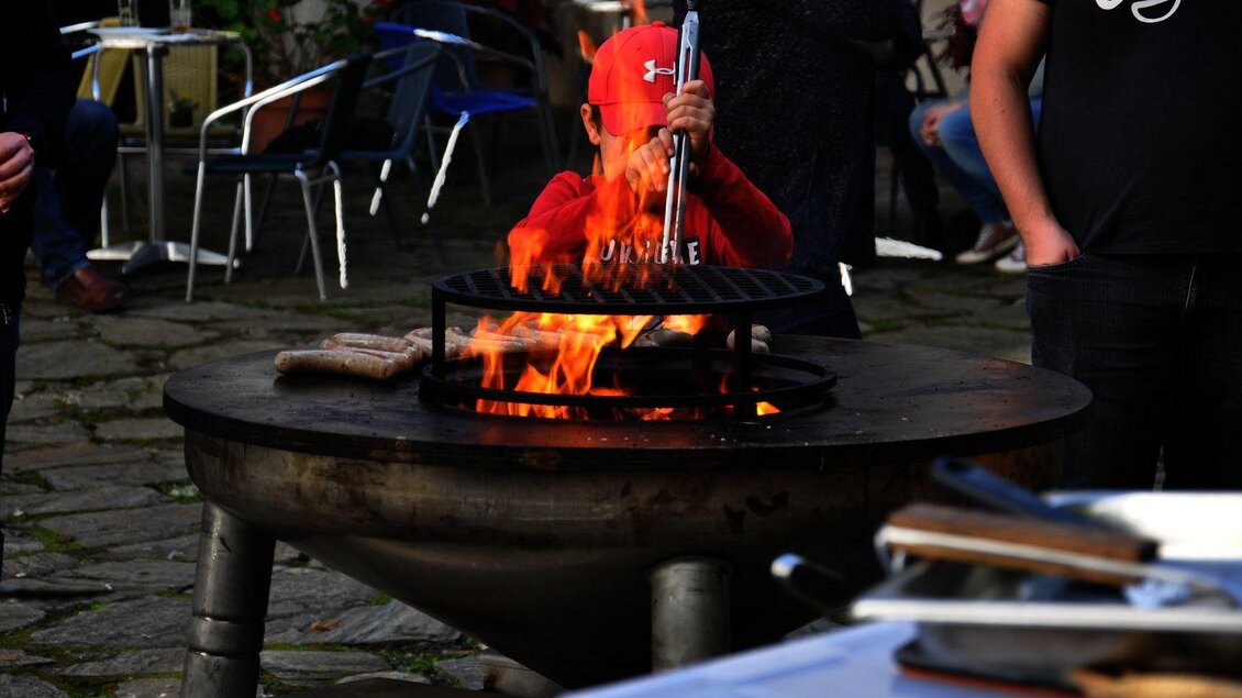Ein Kind in rotem Hut steht an einem Grill, während Flammen aufsteigen. Um ihn herum sind einige Menschen und Tische im Freien.