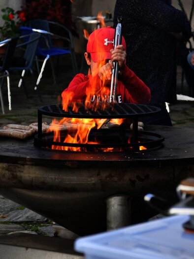 A child in a red hat is standing by a grill as flames rise. Around him are some people and tables outdoors.