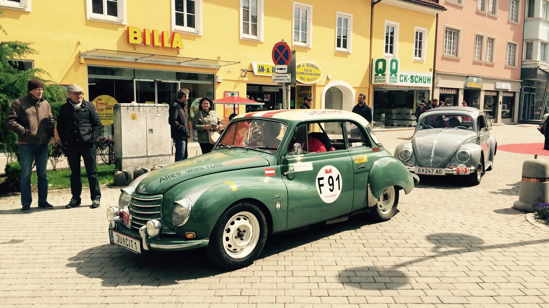 Ein grünes Oldtimer-Auto steht auf einer gepflasterten Straße, während Menschen in der Nähe stehen. Im Hintergrund sind bunte Gebäude und Geschäfte zu sehen. | © FHF Murtal Classic