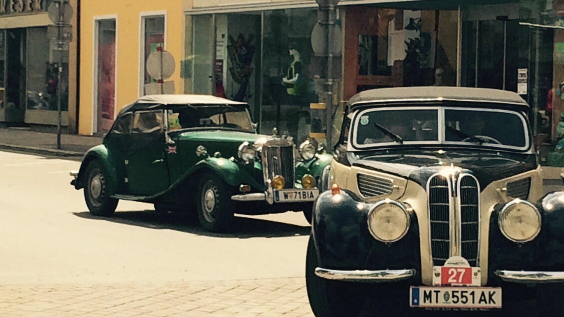 Zwei klassische Autos stehen auf einer Stadtstraße. Im Hintergrund sind Geschäfte und ein klarer Himmel zu sehen. | © FHF Murtal Classic