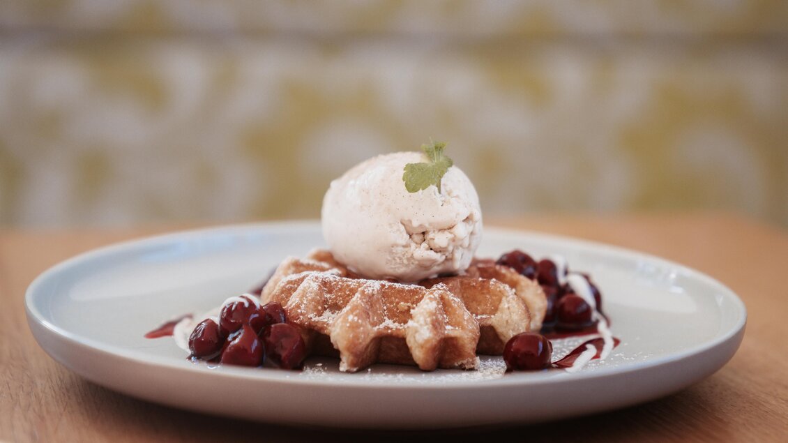 Ein frisch gebackenes Waffle mit einer Portion Eiscreme und roten Früchten. Das Gericht ist dekorativ mit Puderzucker und Sauce angerichtet. | © Martinhof