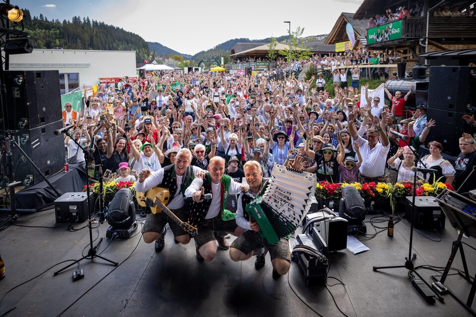 Ein lebhaftes Festival mit einer großen Menge von feiernden Menschen. Auf der Bühne posieren drei Musiker mit Instrumenten und lächeln in die Kamera. | © Edlseer Alm BePo