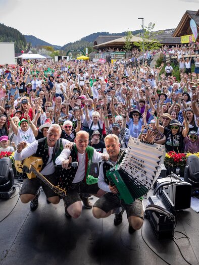 A lively festival with a large crowd of celebrating people. On stage, three musicians pose with instruments and smile at the camera. | © Edlseer Alm BePo