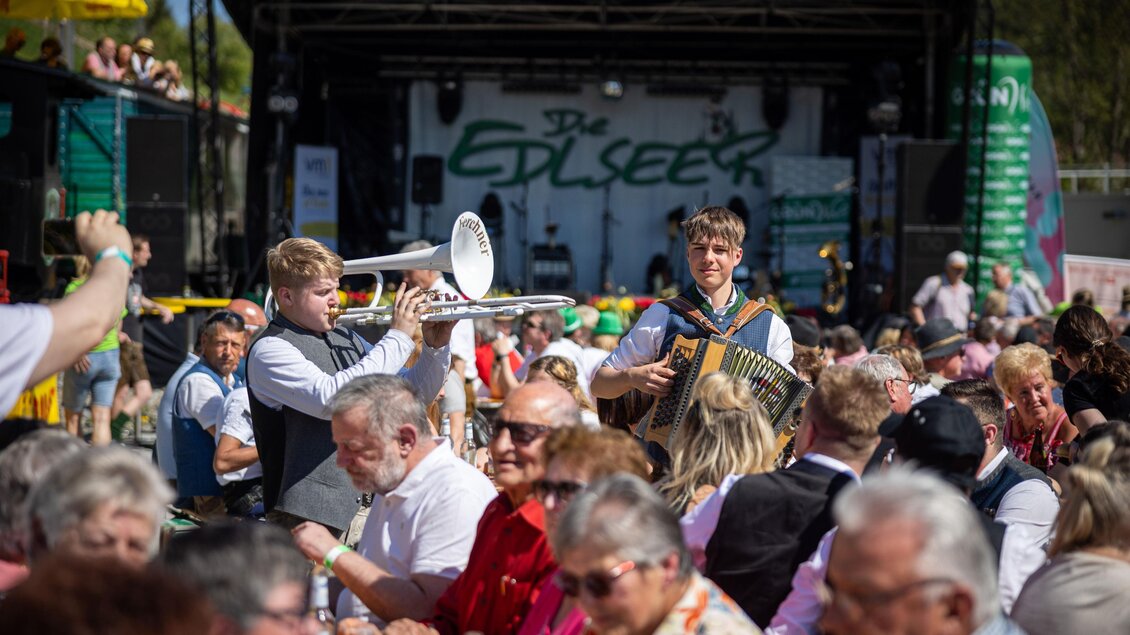 Eine lebhafte Veranstaltung mit Menschen, die an Tischen sitzen und einem Musikperformance zuschauen. Zwei Musiker spielen auf der Bühne und sorgen für gute Stimmung. | © Edlseer Alm BePo