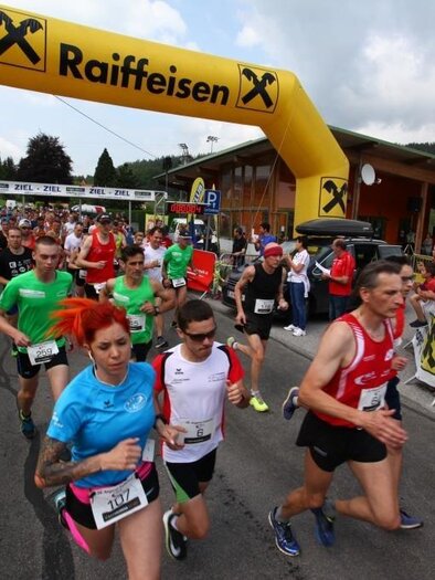 Eine Gruppe von Läufern startet einen Wettlauf unter einem auffälligen Startbogen. Die Teilnehmer tragen bunte Sportkleidung und die Stimmung ist enthusiastisch. | © Happy Lauf Anger