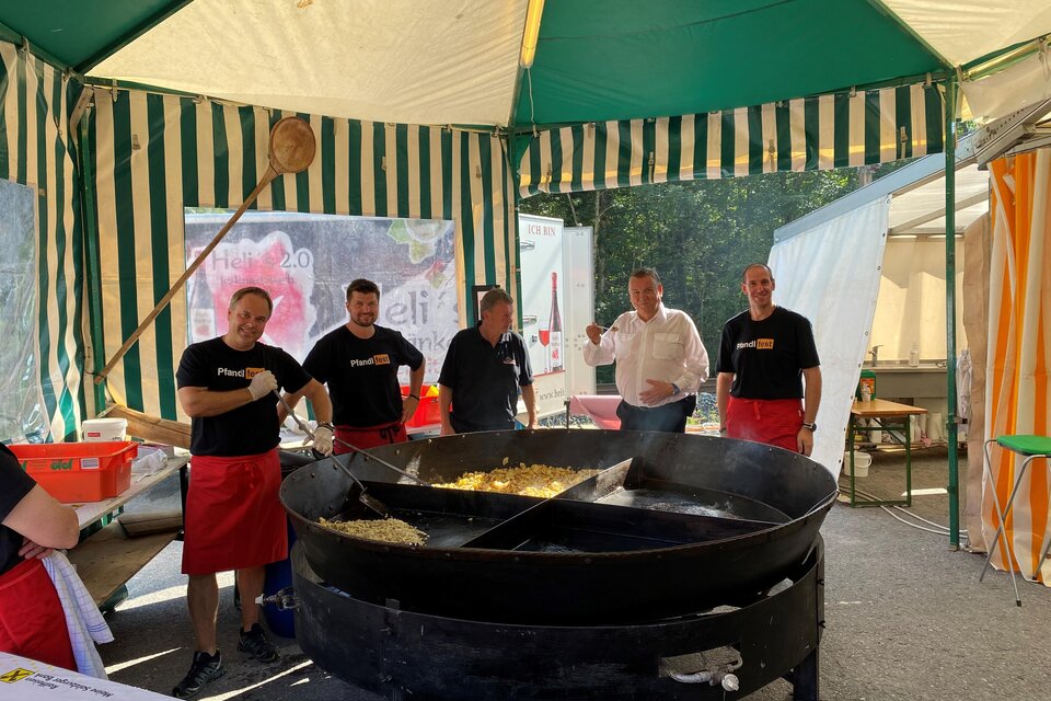 Ein festlich dekorierter Zeltstand mit mehreren Männern in schwarzen T-Shirts und roten Schürzen, die an einem großen Kessel mit Essen arbeiten. Im Hintergrund sind Zeltwände und andere Stände sichtbar. | © FF Mandling