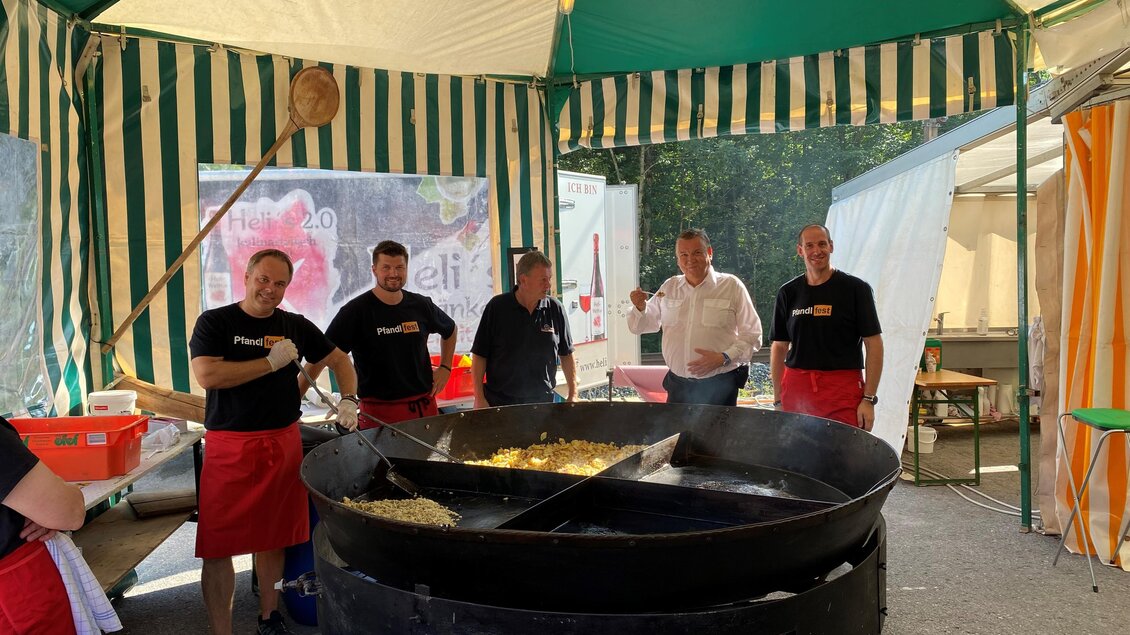 Ein festlich dekorierter Zeltstand mit mehreren Männern in schwarzen T-Shirts und roten Schürzen, die an einem großen Kessel mit Essen arbeiten. Im Hintergrund sind Zeltwände und andere Stände sichtbar. | © FF Mandling