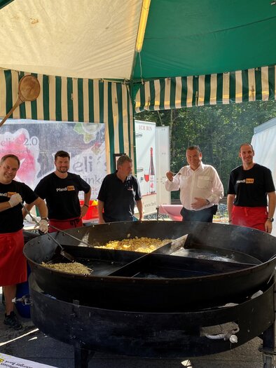 A festively decorated tent stand with several men in black T-shirts and red aprons working at a large cauldron of food. In the background, tent walls and other stands are visible. | © FF Mandling