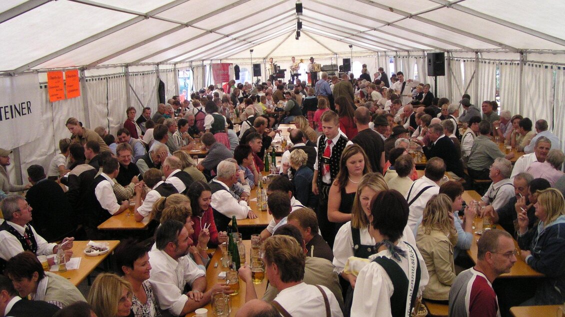 Ein festliches Zelt voller Menschen, die an langen Tischen sitzen und feiern. Die Gäste tragen traditionelle Kleidung und genießen Essen und Getränke. | © FF Mandling